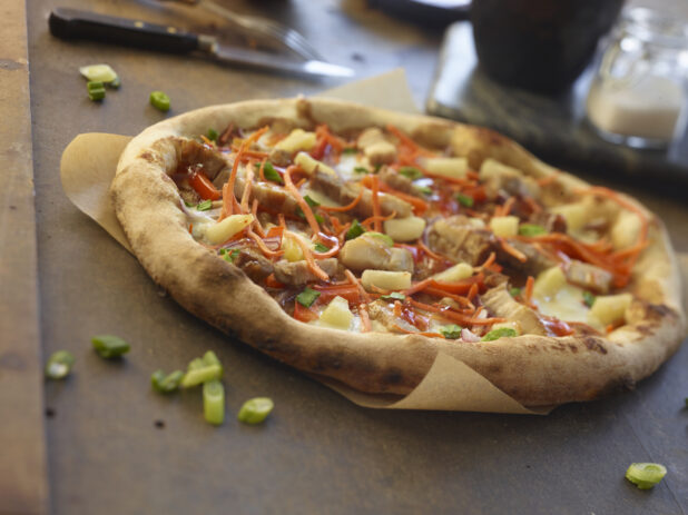 Whole pizza with pork belly, pineapple, shredded carrots, red peppers and green onions on parchment paper, close-up