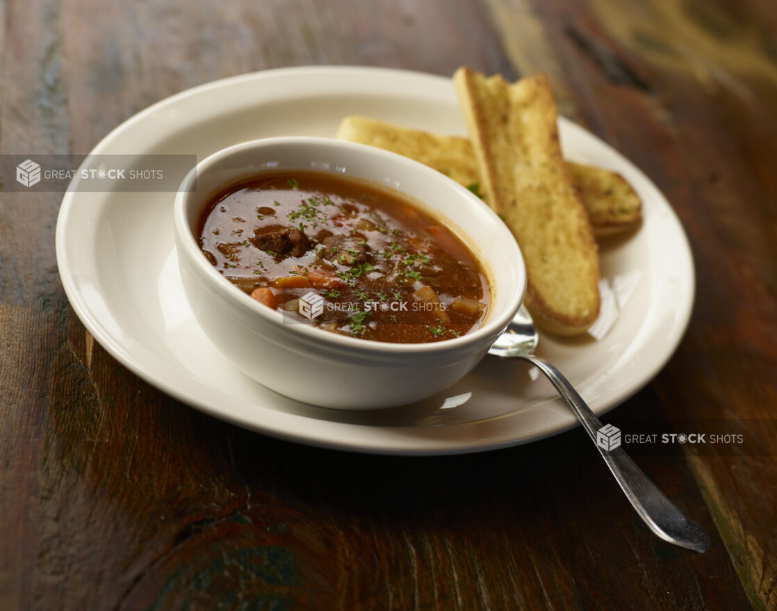 Minestrone soup in a white bowl on a white plate with garlic toast on the side, dark wood background