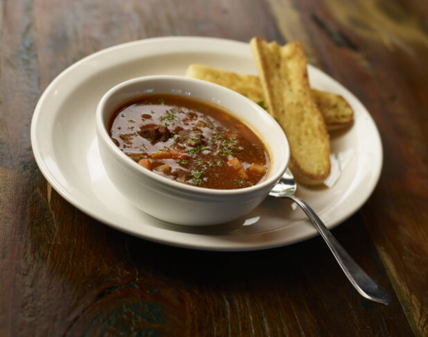 Minestrone soup in a white bowl on a white plate with garlic toast on the side, dark wood background