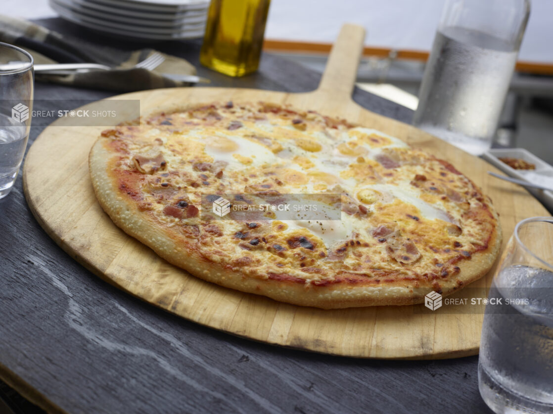 Whole breakfast pizza with bacon and eggs on a round wood pizza peel, grey wood background, close-up, tilted angle