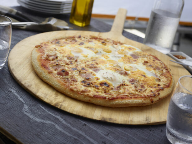 Whole breakfast pizza with bacon and eggs on a round wood pizza peel, grey wood background, close-up, tilted angle