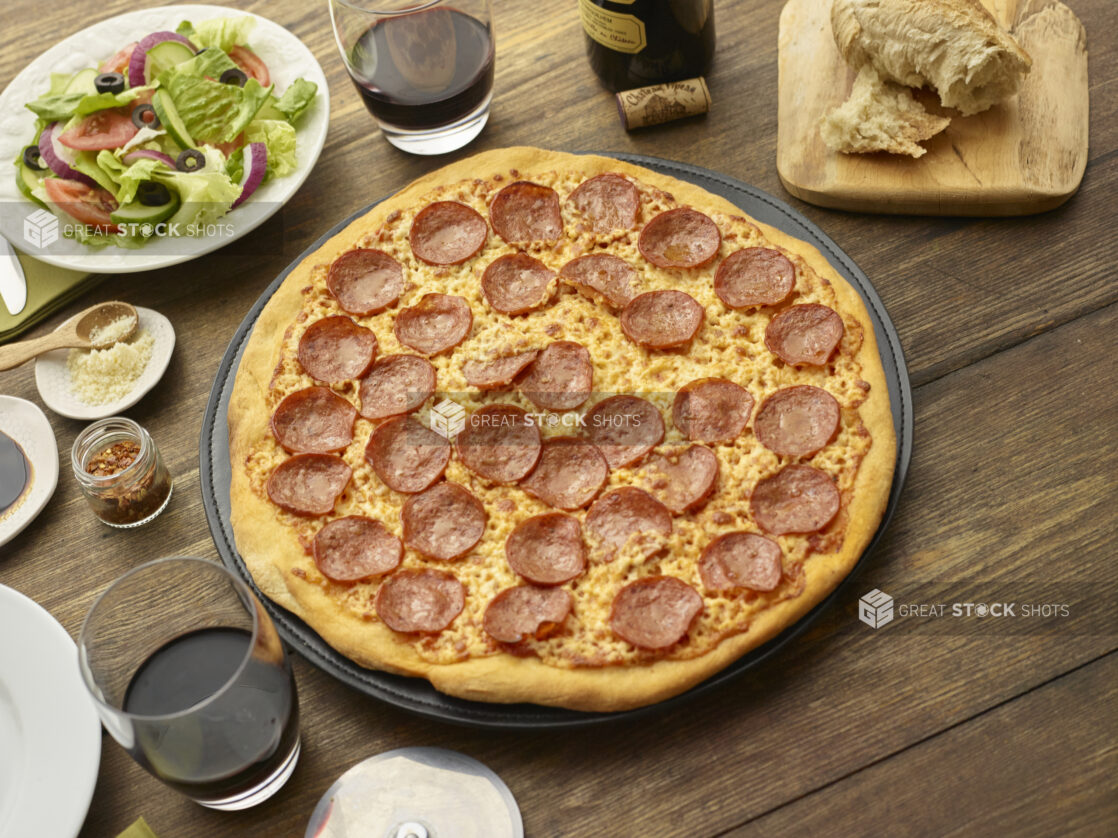 Whole unsliced pepperoni pizza surrounded by tumblers of red wine, torn Italian bread, side salad and condiments, wood background