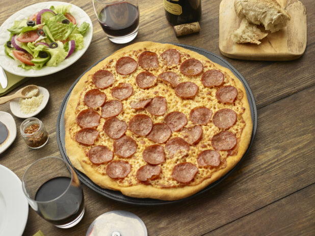 Whole unsliced pepperoni pizza surrounded by tumblers of red wine, torn Italian bread, side salad and condiments, wood background