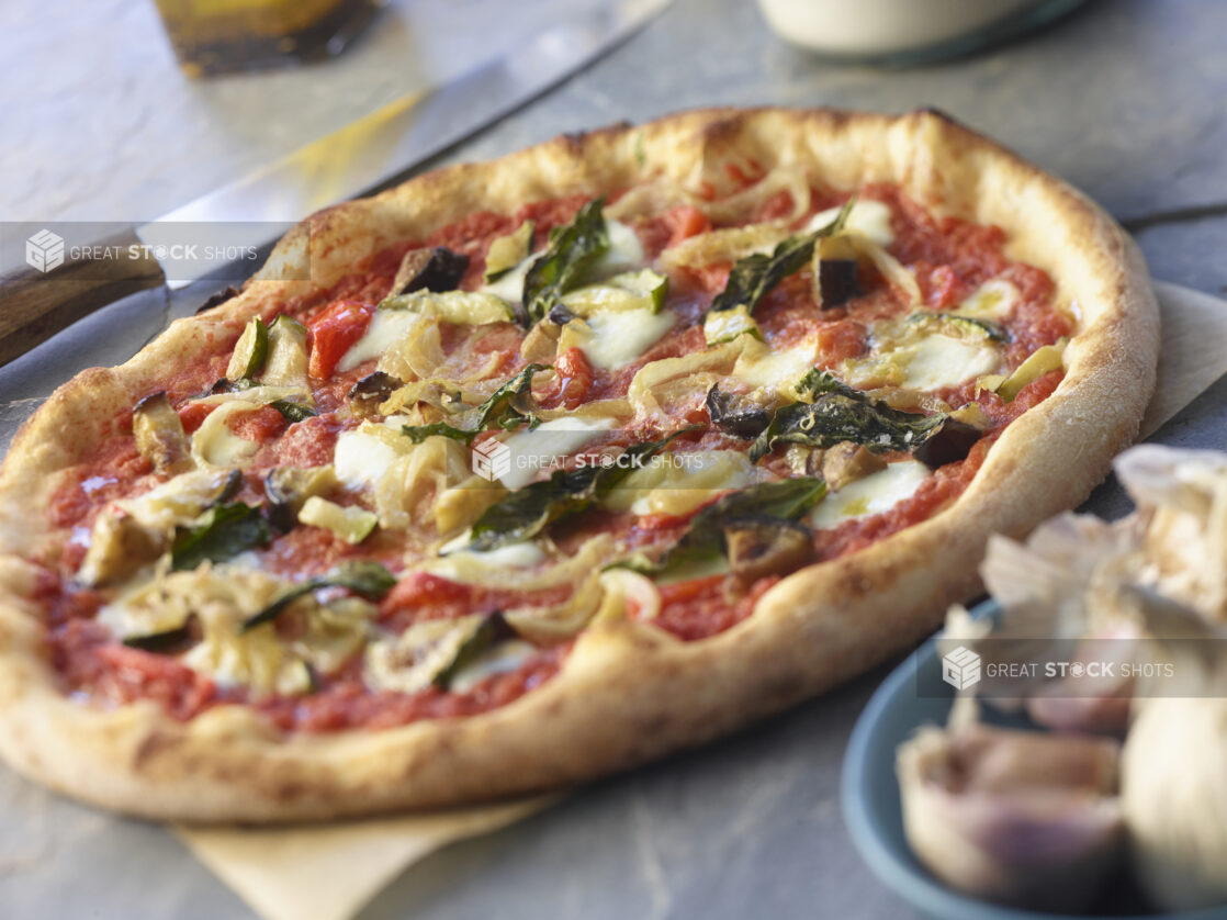 Whole 3 topping pizza with bocconcini, artichokes, eggplant, and basil on parchment paper, knife in background close-up