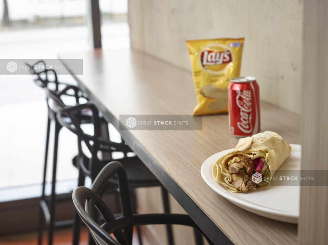 Middle eastern shawarma pita on a plate with a can of coke and bag of plain chips in a restaurant on a wooden table