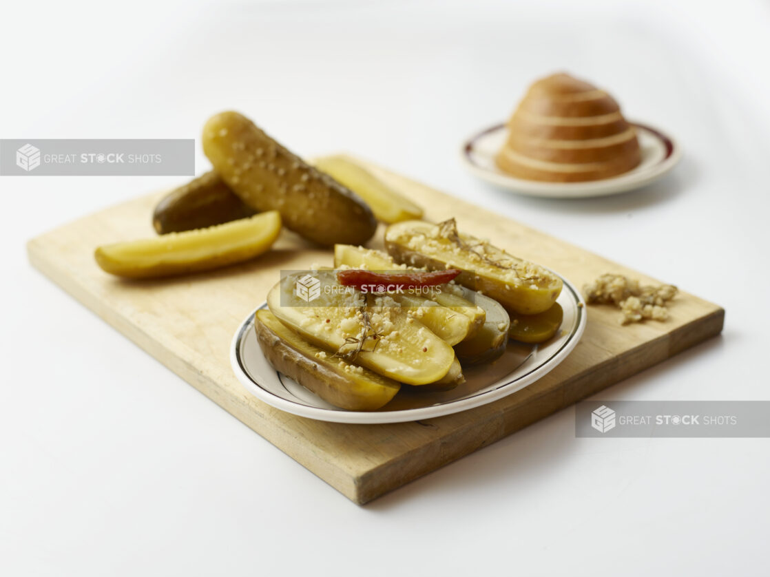 Side plate piled with halved dill pickles garnished with roasted garlic with more pickles piled behind, all on wood board, white background