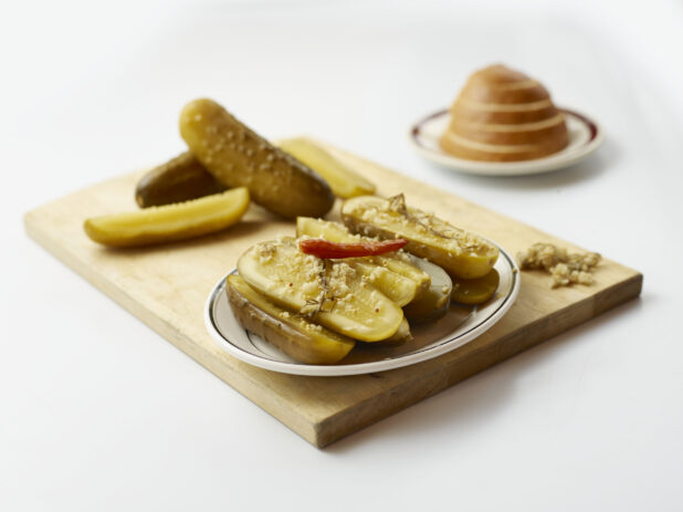 Side plate piled with halved dill pickles garnished with roasted garlic with more pickles piled behind, all on wood board, white background