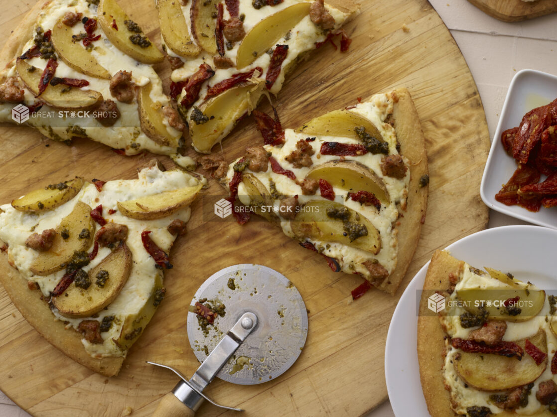 Sliced pizza with potato wedges, sundried tomatoes, and sausage on a wooden pizza peel with cutter, close-up, overhead view