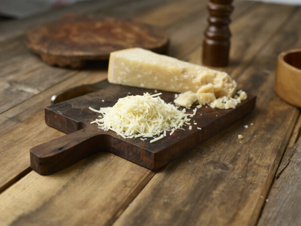 Parmesan cheese wedge, partially crumbled and shredded, on a wood cheese board, wood background