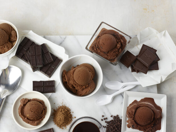 Many bowls of chocolate ice cream surrounded by chocolate and spoons