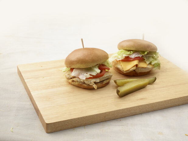 Two grilled chicken burgers, one with cheese and one without, and two dill pickle spears on a wood board, white background