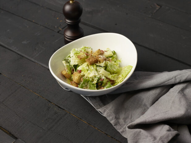 Side Caesar salad in a small white ceramic bowl, pepper grinder and cloth napkin alongside, black wood background