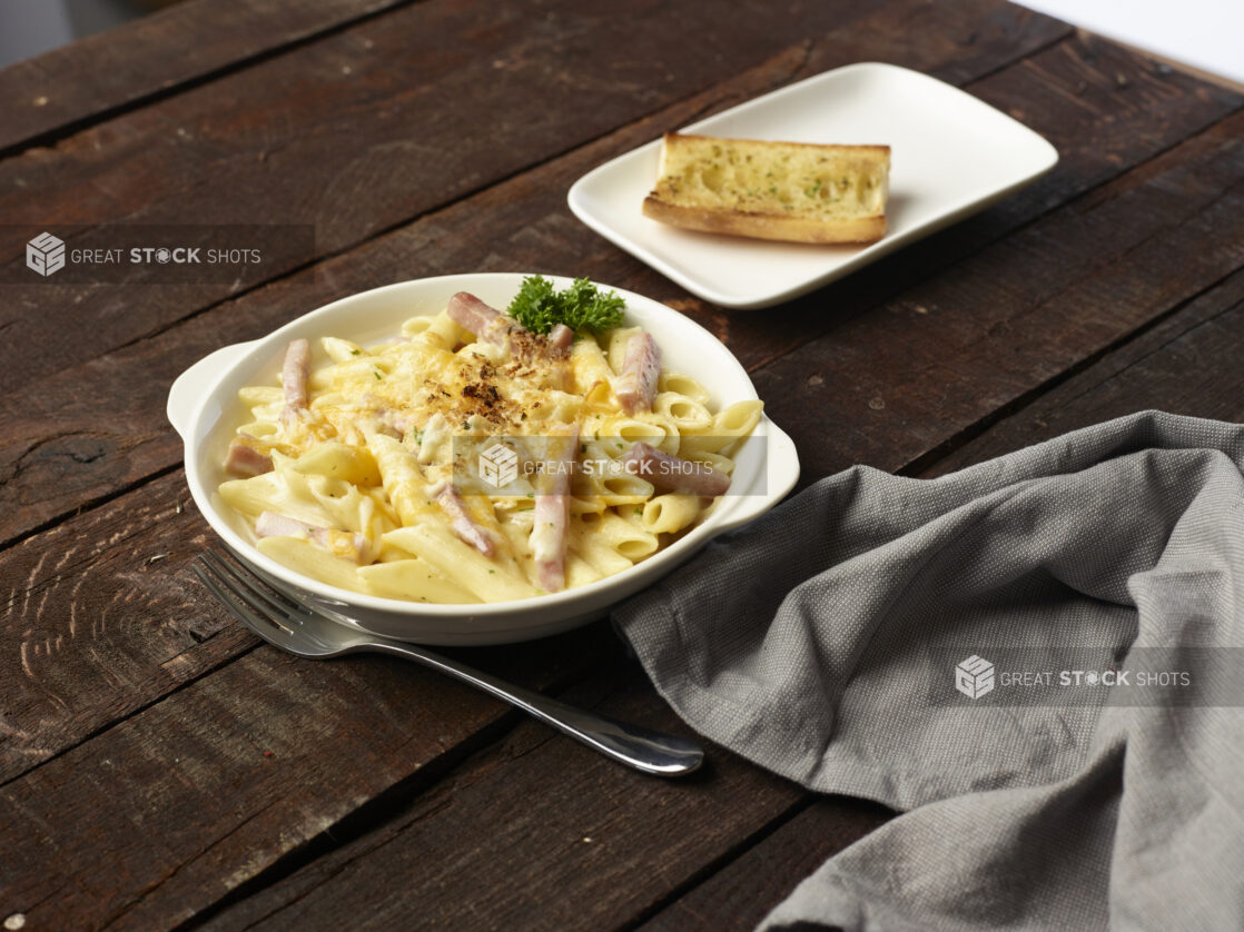 Penne Alfredo with pork belly in a white bowl with handles, side of garlic bread on white plate in background, dark wood background