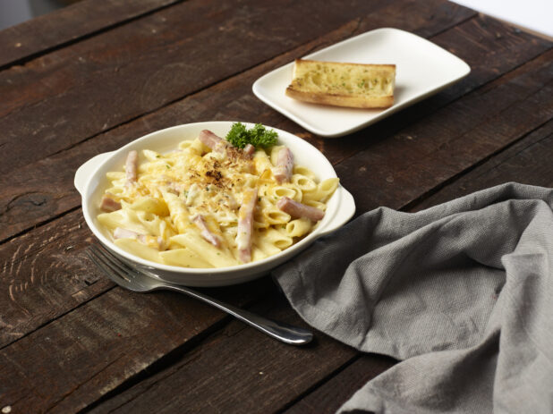 Penne Alfredo with pork belly in a white bowl with handles, side of garlic bread on white plate in background, dark wood background