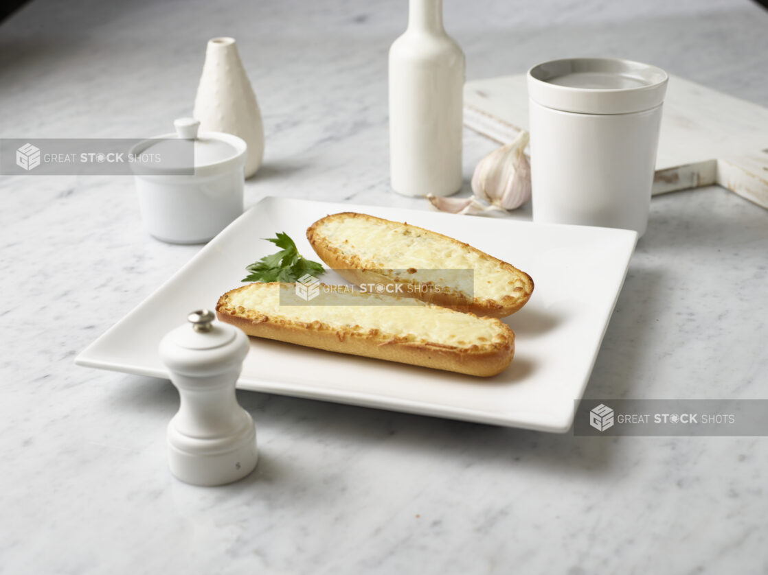 Stick of garlic bread, open-faced, with melted mozzarella cheese, on a square white plate, white kitchenware surrounding, white marble background