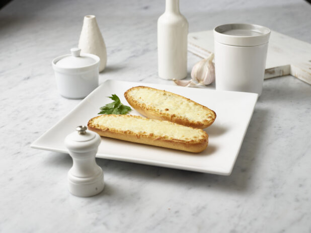 Stick of garlic bread, open-faced, with melted mozzarella cheese, on a square white plate, white kitchenware surrounding, white marble background