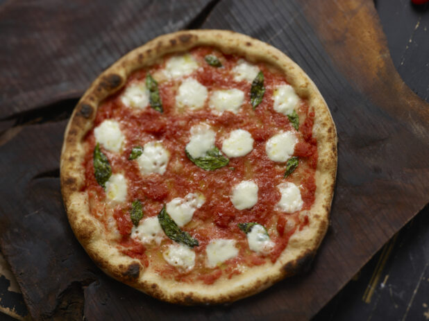 Whole margherita pizza overhead with fresh basil on a dark wooden board