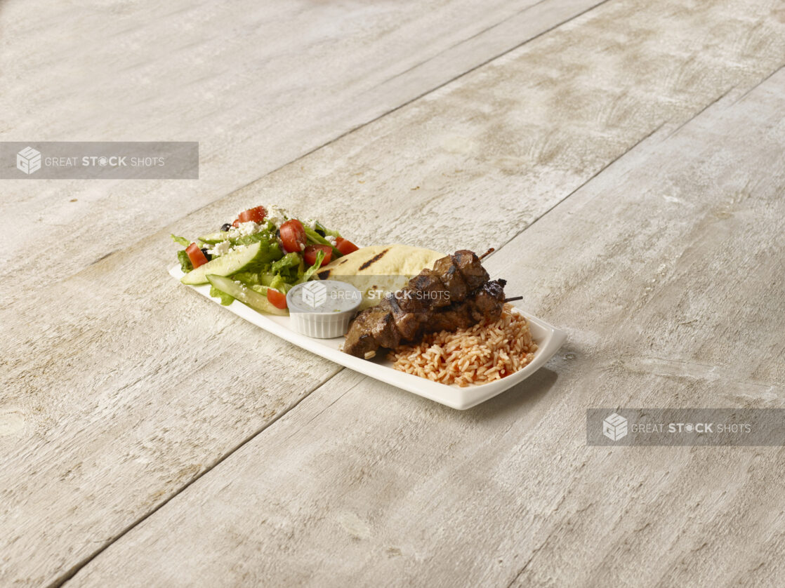 Beef kebabs on a bed of rice, greek salad and pita with creamy dip in a rectangular white plate on a wooden background