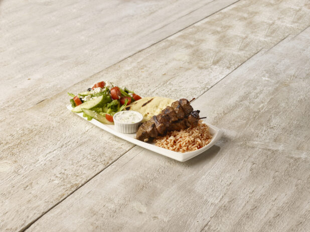 Beef kebabs on a bed of rice, greek salad and pita with creamy dip in a rectangular white plate on a wooden background