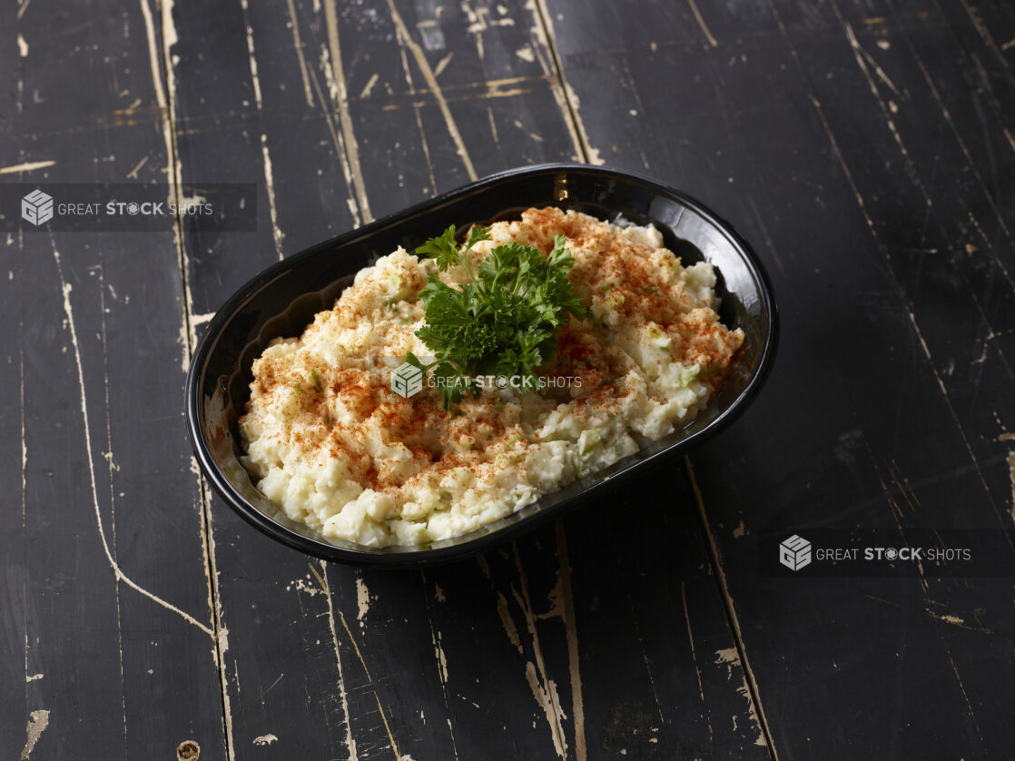 Black bowl of potato salad topped with paprika and parsley on a distressed black wooden background