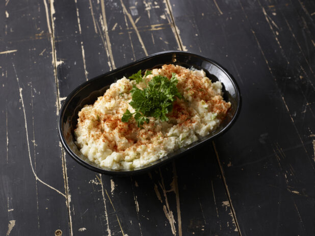 Black bowl of potato salad topped with paprika and parsley on a distressed black wooden background