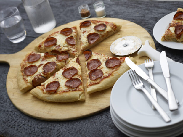 Sliced small pepperoni pizza with slice missing on a pizza peel with a stack of plates and cutlery