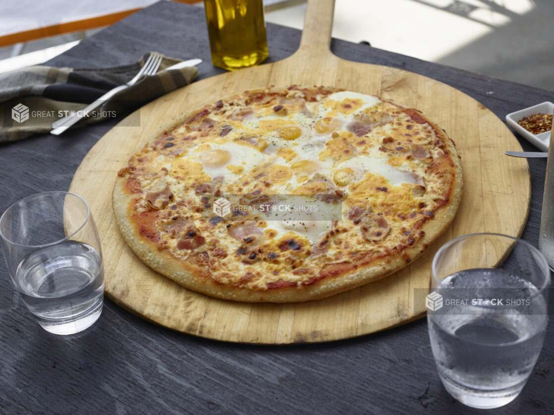 Whole breakfast pizza with eggs and bacon on a pizza peel on a black wooden table