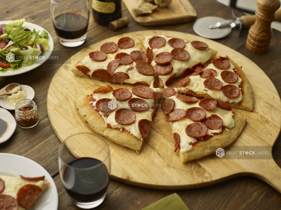 Sliced small pepperoni pizza with slice removed on a pizza peel with a side salad and red wine glasses