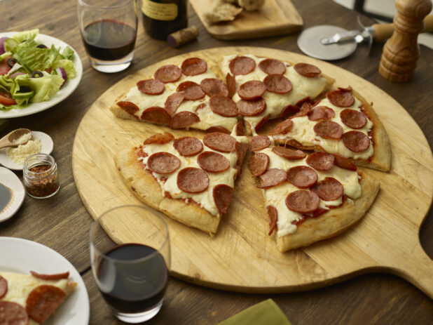 Sliced small pepperoni pizza with slice removed on a pizza peel with a side salad and red wine glasses