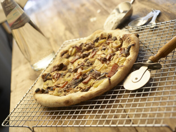 Meat bacon pizza on a cooling rack beside pizza cutting wheel