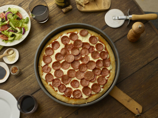 Overhead view of a full deep dish pepperoni pizza surrounded by red wine glasses and a side salad