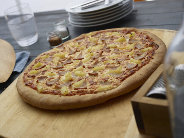 Whole Hawaiian pizza on a wooden board with a stack of white plates in the background