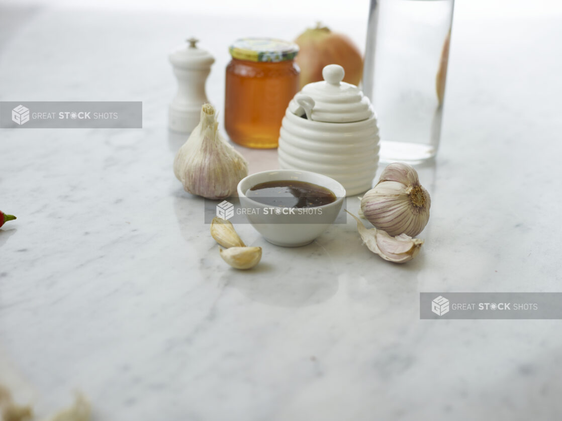 Small white bowl of honey garlic sauce surrounded by ingredients on white marble