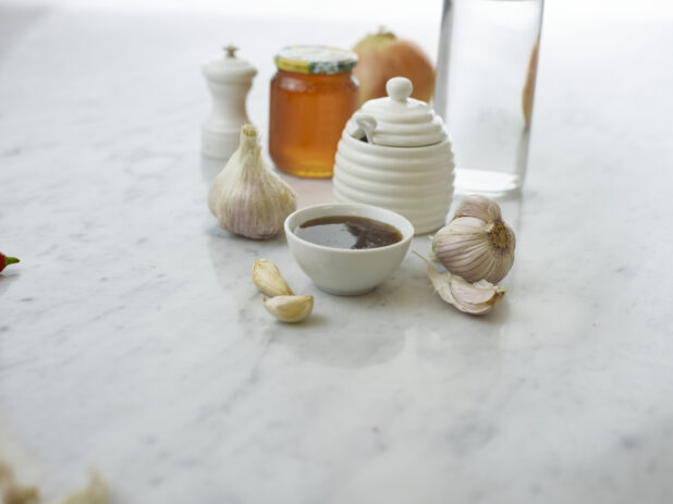 Small white bowl of honey garlic sauce surrounded by ingredients on white marble