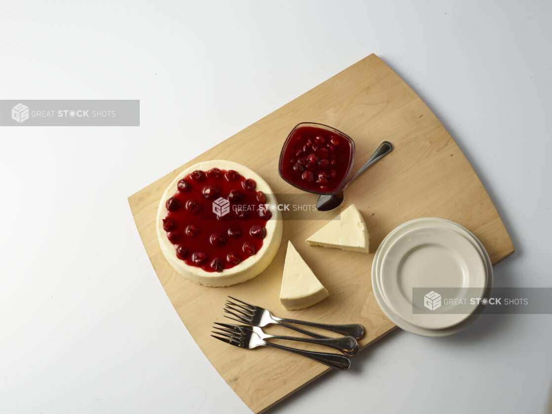 Overhead of small cherry cheesecake with slices of plain cheese cake on the side and a bowl of cherry compote on a wooden board