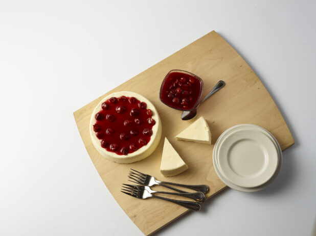 Overhead of small cherry cheesecake with slices of plain cheese cake on the side and a bowl of cherry compote on a wooden board