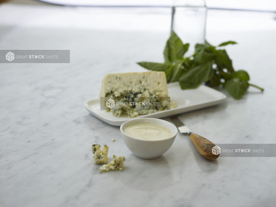 Small white bowl of blue cheese dip surrounded by ingredients on white marble