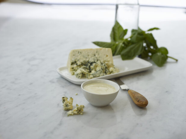 Small white bowl of blue cheese dip surrounded by ingredients on white marble