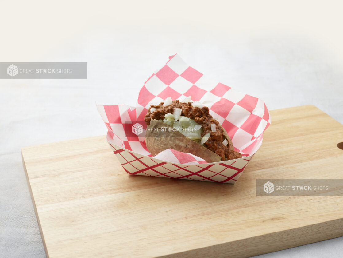 Loaded baked potato with beef chili and onions in a red and white cardboard basket on a wooden board