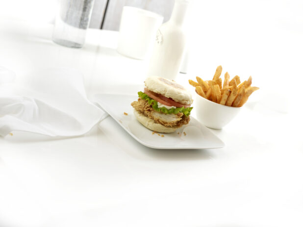 Fried chicken breakfast sandwich on a white plate with a side of french fries on a white background
