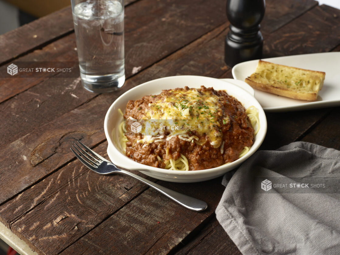 Spaghetti topped with melted cheese in a white baking dish with garlic bread on a wooden table