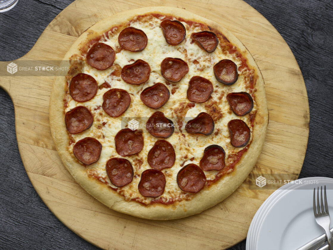 Overhead view of a full pepperoni pizza on a wooden peel on a black wooden background