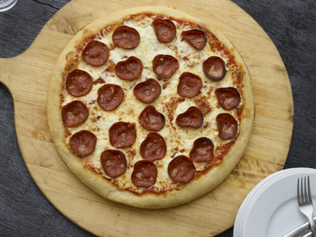 Overhead view of a full pepperoni pizza on a wooden peel on a black wooden background
