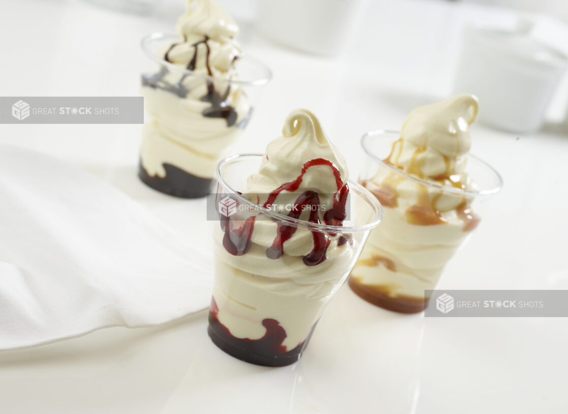 Three vanilla sundae cups with different flavors, chocolate, strawberry and caramel, on a white counter