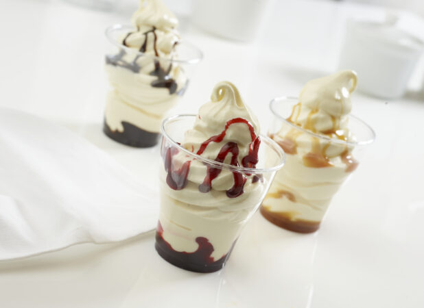 Three vanilla sundae cups with different flavors, chocolate, strawberry and caramel, on a white counter