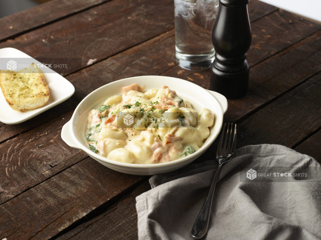 Baking dish of creamy gnocchi with a side of garlic bread and cloth napkin on a dark wooden background