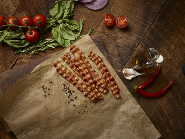 Strips of crispy bacon on parchment surrounded buy fresh ingredients on a dark wood background