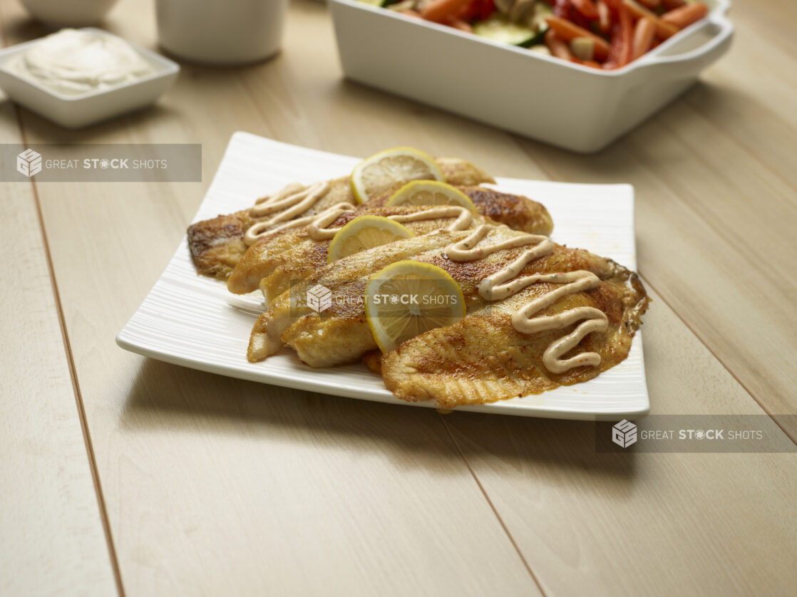 Seasoned white fish fillets on a white plate garnished with lemon slices and drizzled with a creamy sauce, wood background