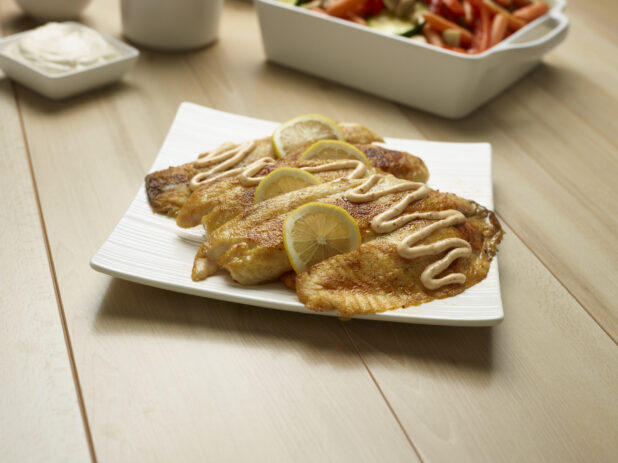 Seasoned white fish fillets on a white plate garnished with lemon slices and drizzled with a creamy sauce, wood background