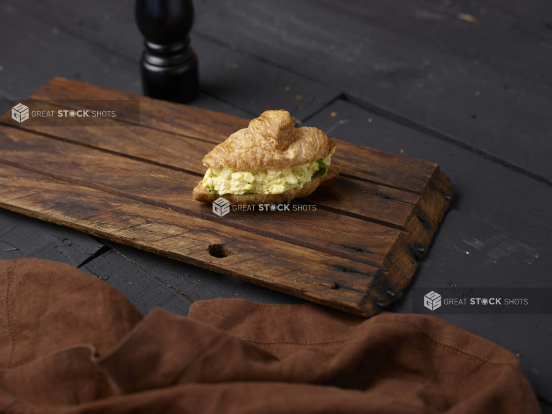 Chopped egg salad croissant sandwich with lettuce on a wooden board with brown cloth napkin